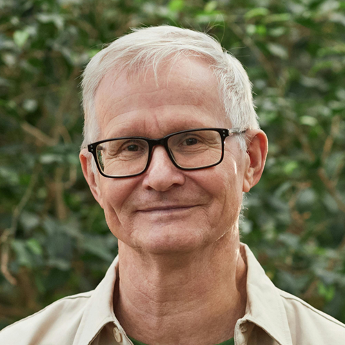 Smiling older man wearing glasses, standing outdoors in front of greenery.